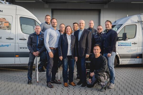 Das Team eines Handwerksbetriebs steht lächelnd vor Firmenfahrzeugen auf dem Betriebshof.