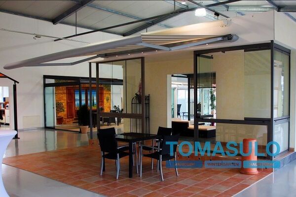 Moderner Ausstellungsraum mit verglastem Wintergarten, Markise und Sitzgruppe innen.