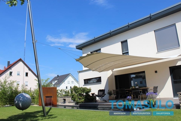 Modernes Einfamilienhaus mit Sonnensegel, Terrasse und Garten bei blauem Himmel.