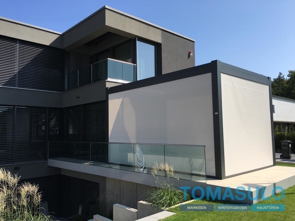 Moderne Terrasse mit Glasgeländer und Pergola-Markise an einem Einfamilienhaus bei Sonne.